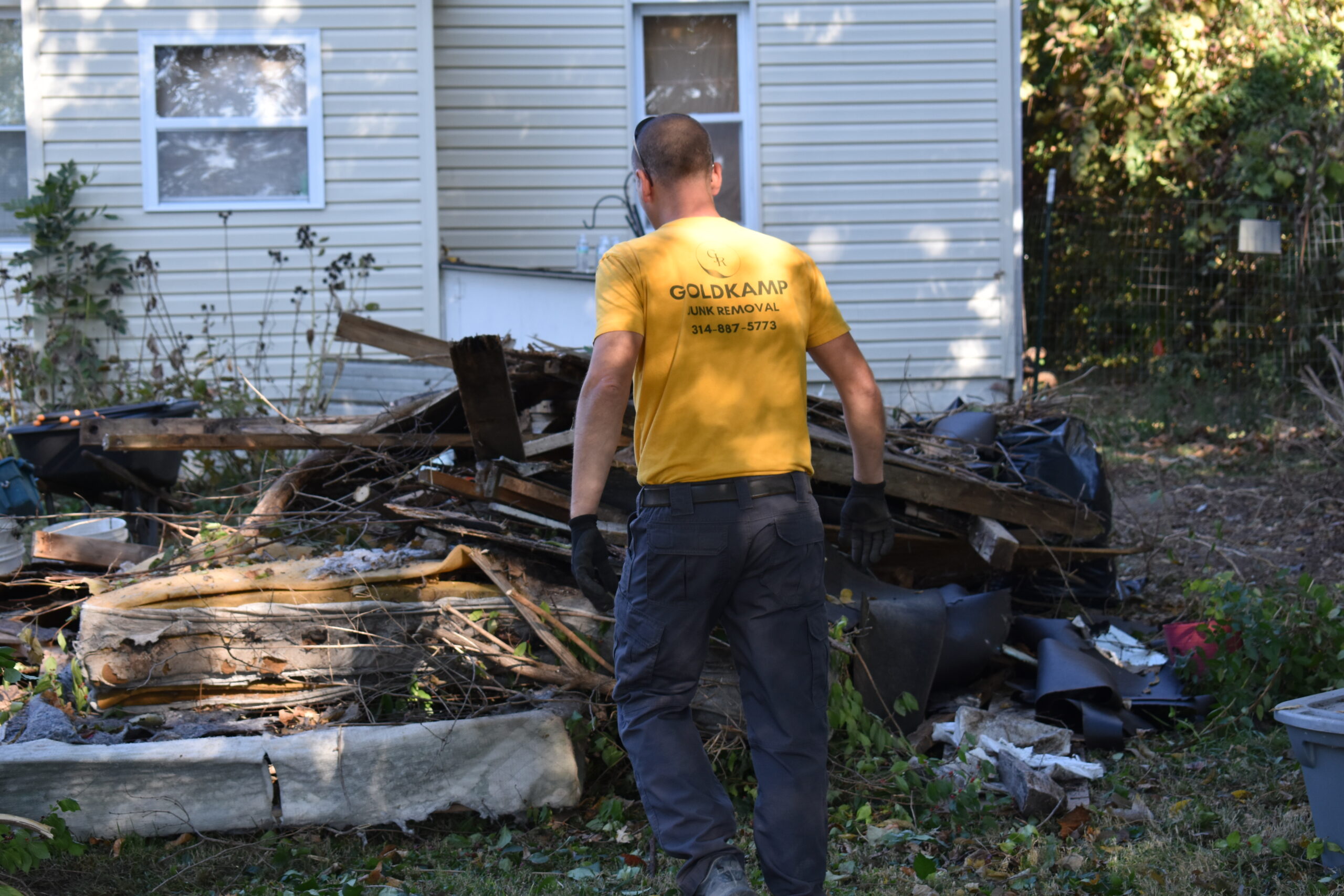 Goldkamp Junk Removal worker facing a large pile of wooden debris and junk from a demolished shed, with a house in the background.