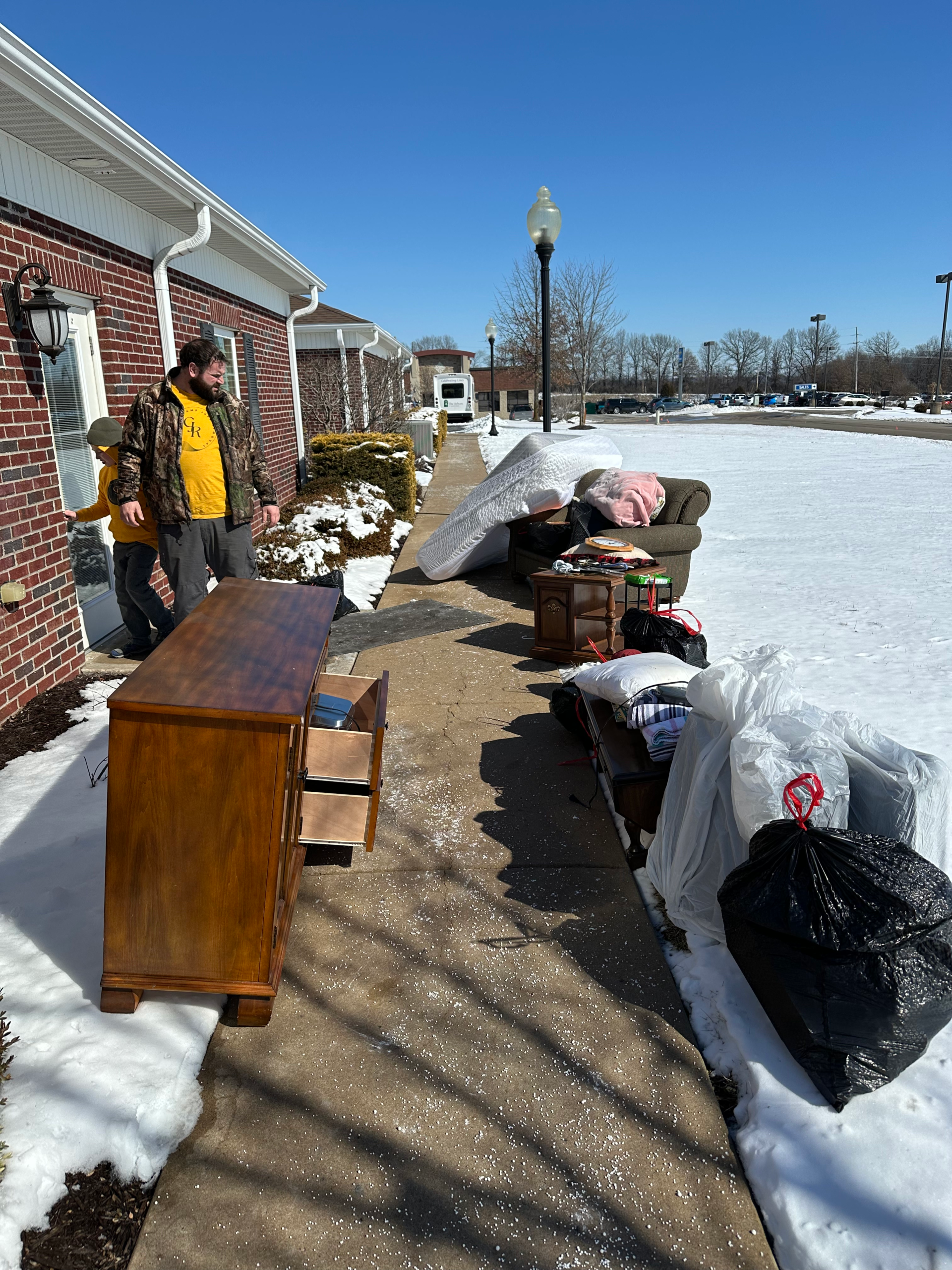 Along the sidewalk are various discarded furniture items being hauled away, including a wooden dresser, couch, mattress, nightstand, and bags of trash, with a moving truck parked in the background. The service depicted is furniture hauling.