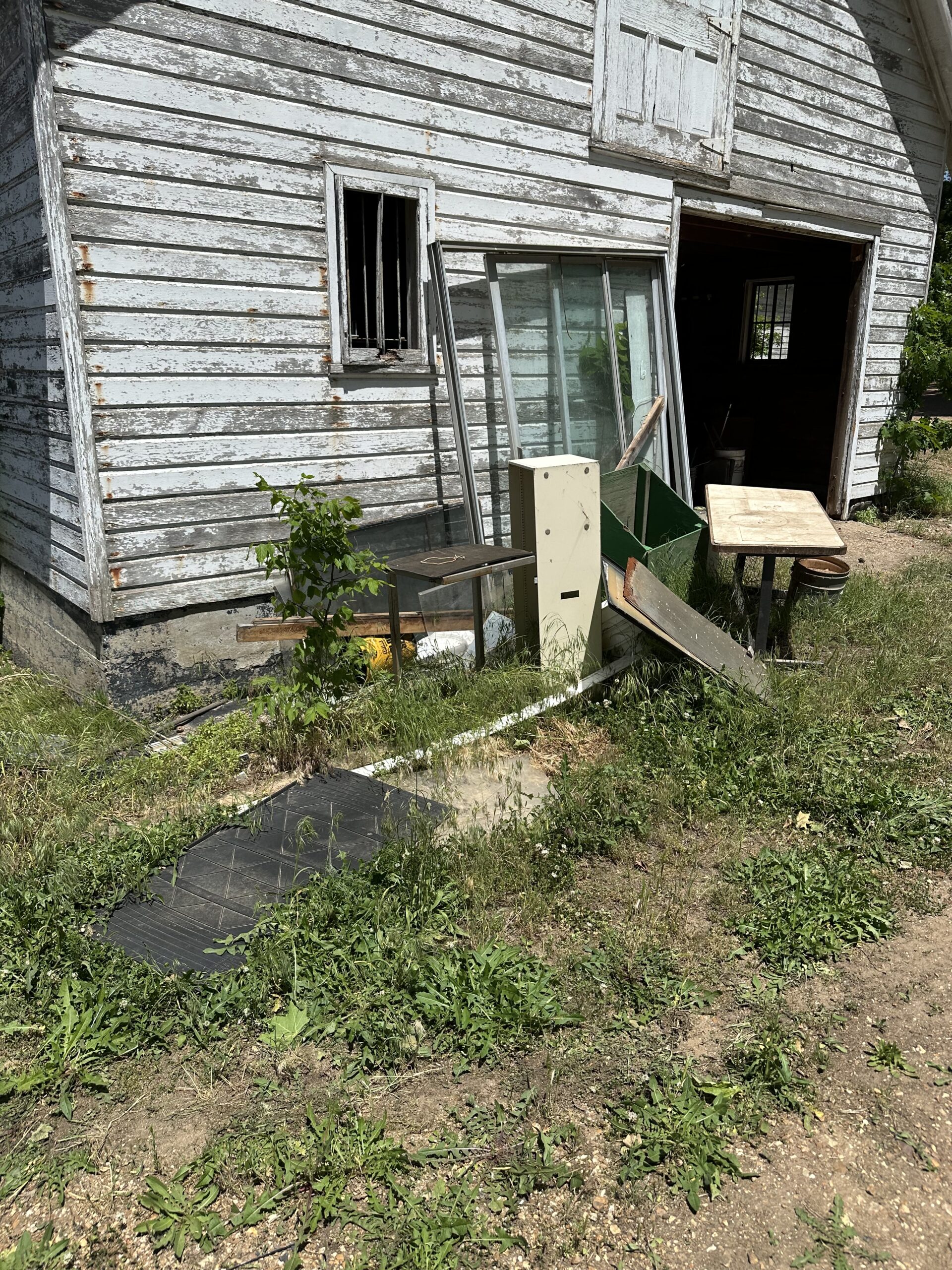 Yard waste removal in O'Fallon, MO: Overgrown weeds and miscellaneous debris around a weathered white building, showing an area in need of professional yard clean-up.