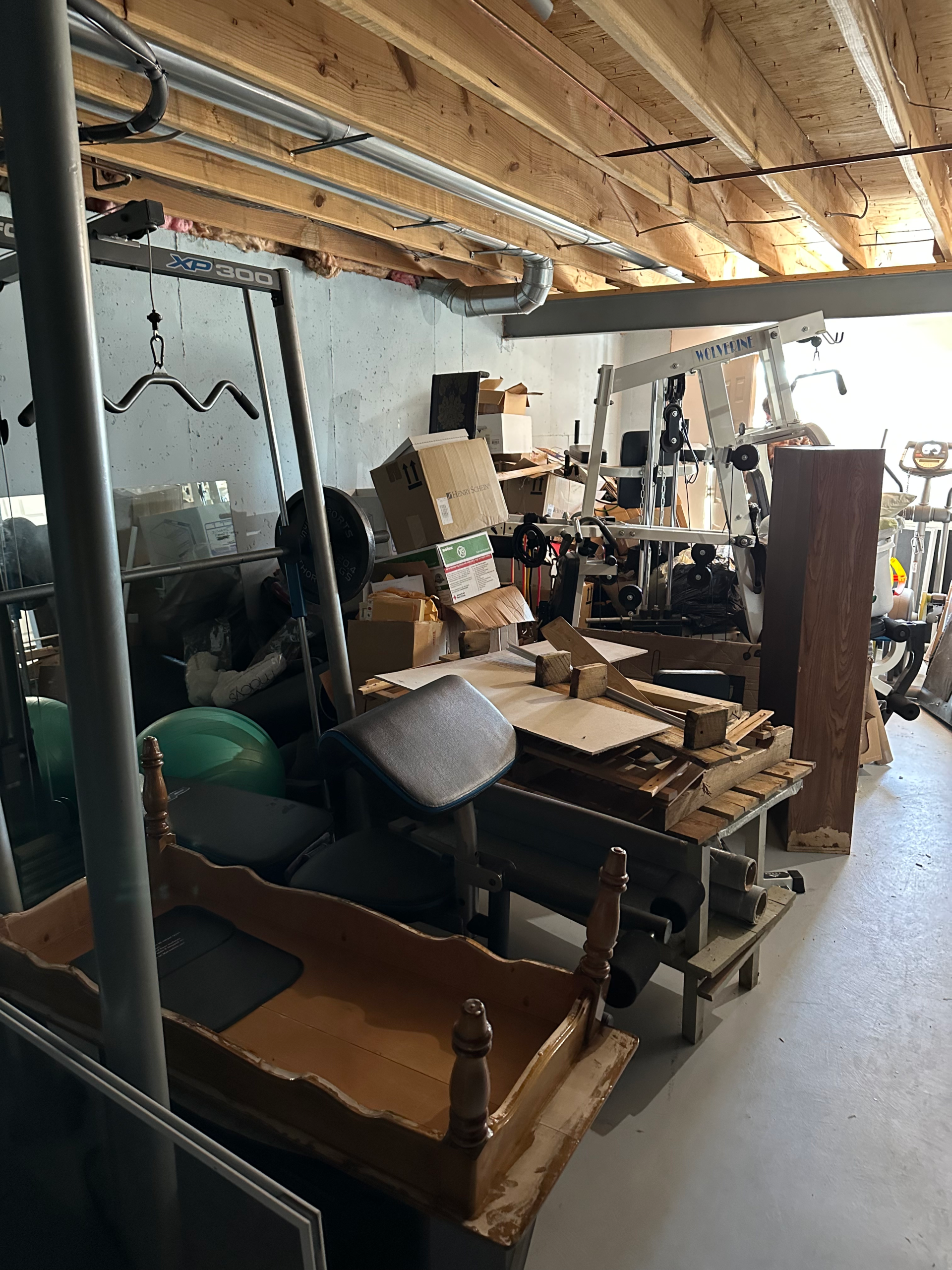 A cluttered basement scene showing mixed storage of workout equipment (cable machine labeled “XP 300”), cardboard boxes, wooden furniture pieces (including a partially stripped drawer), exercise balls, and miscellaneous lumber piled on a workbench, ready for furniture hauling service.