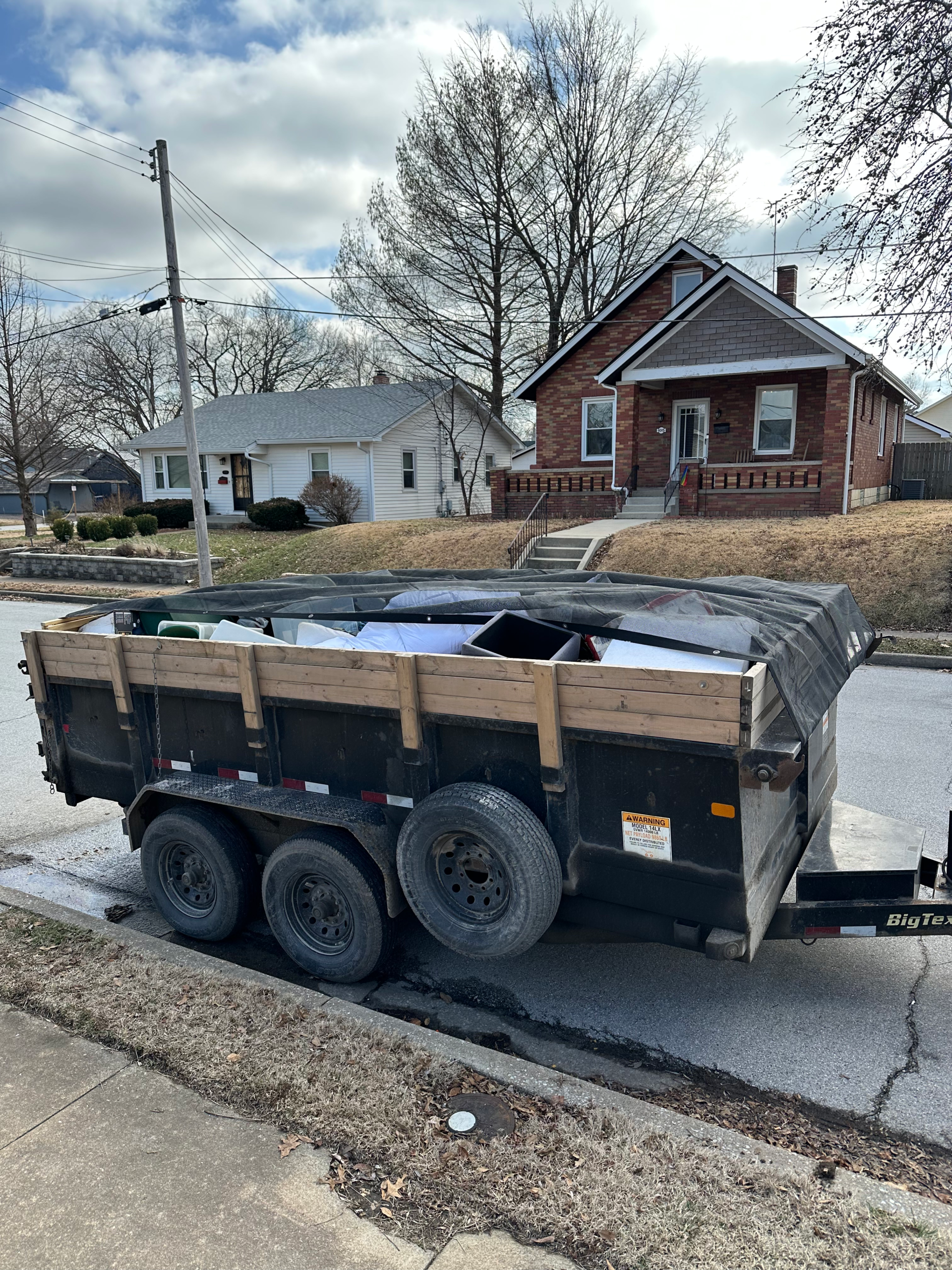 black utility trailer filled with various scrap metal pieces and other debris, covered with a mesh tarp, parked on a residential street in front of a brick house, ready for scrap metal removal.